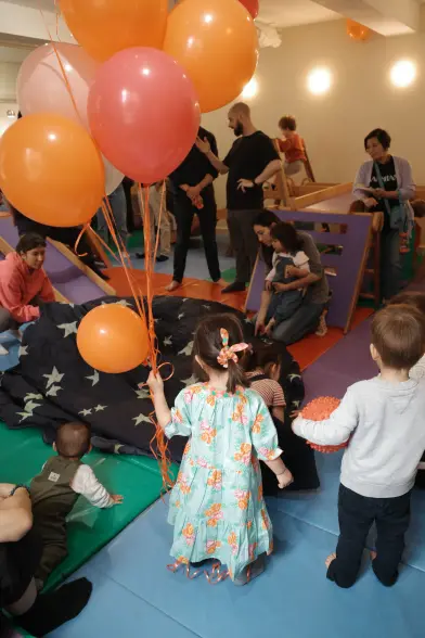 a ruckenfigure portrait of my daughter, a 3yo in a flowery dress, holding a bunch of floating balloons inside a softplay venue, two kids by her sides with more kids and parents in the background