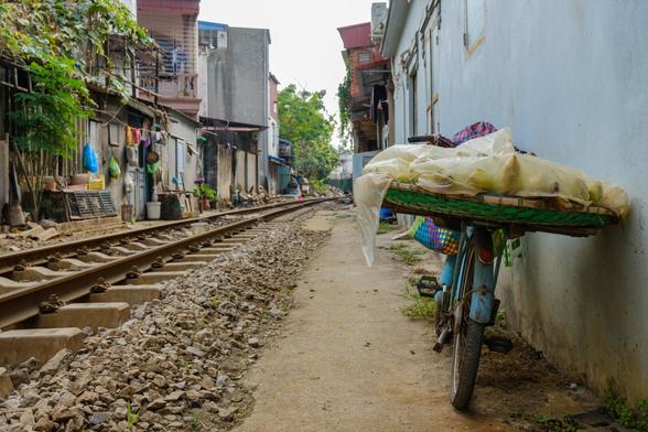Ein schmaler Weg neben einem Bahngeis wird von Wohngebäuden flankiert. Im Vordergrund hält ein blaues Fahrrad einen gewebten Korb, der mit einer Plastikfolie bedeckt ist, unter der gerade so eben Bananen zu erkennen sind. Die umliegende Vegetation und Trümmer sind entlang des Weges sichtbar.