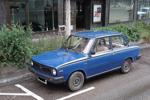 Ein blauer Volvo mit H im Nummernschild parkt auf einem markierten Parkstreifen am Straßenrand Die Front ist links, man sieht die linke Seite, das Heck ist rechts.
Hinter dem Wagen ist ein Grünstreifen, dahinter Gehweg und Gebäude.