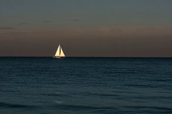 Die untere Hälfte des Bildes zeigt das blaue, ruhige Wasser der Ostsee, hier in der Flensburger Förde. Am Horizont ist ein Segelschiff mit weißen Segeln zu sehen. Darüber spannt sich der Himmel mit dem Orangerot der aufgehenden Sonne, die noch nicht zu sehen ist, und darüber mit dem Azurblau des Himmels.
