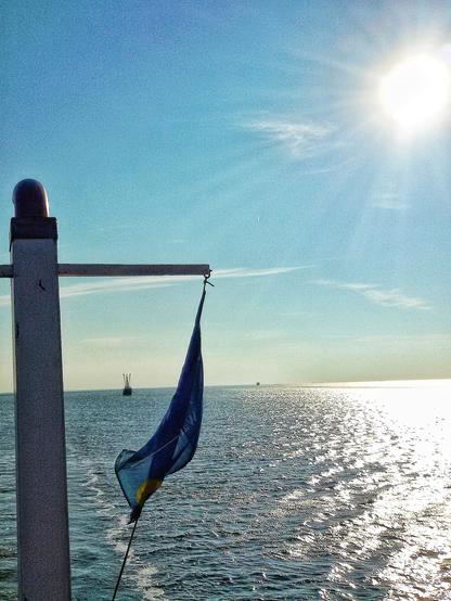 Der Blick von einem Schiff aus geht über das Heck auf die ruhige, offene See. Links ist ein Schiffsmast mit Flagge zu sehen, in der Ferne sind zwei Boote an verschiedenen Positionen erkennbar. Oben rechts im Bild steht die Sonne und wirft ihre Strahlen in den blauen Himmel.