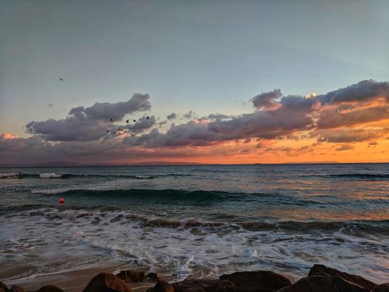 A wide shot captures a stunning sunset over a beach, with an intense orange glow illuminating the horizon and the undersides of scattered clouds. A flock of birds flies in a V-formation across the upper left sky. The sea is a deep blue-green, with frothy white waves breaking on the sandy shore in the foreground. A surfer is visible in the water on the far left, riding a wave. A lone orange buoy floats in the mid-ground of the water. Dark rocks are visible at the very bottom edge of the image.