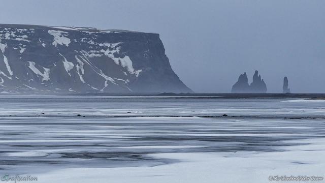 A photo of a frozen landscape which is actually in colour but looks more like monochrome. The foreground is the edge of a lake which has turned to ice, and stripes of clean snow partially cover the dark surface. In the distance a black mountain rises, dark rock with ice on any surface which isn't vertical. On the right are the silhouettes of a pair of sea stacks, one has two distinct points and the other is solitary. The sky is grey and snowy. It's quite bleak.