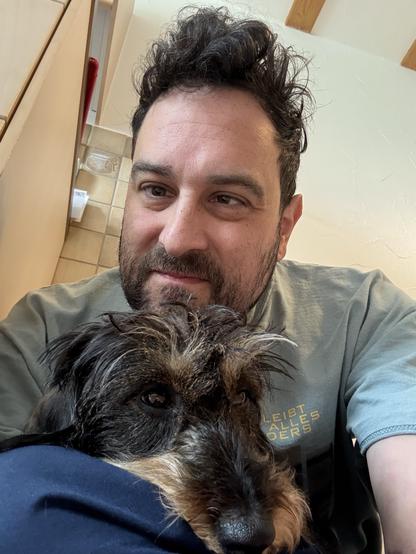 A person with dark curly hair is smiling while sitting next to a small dog with wiry fur. The dog is resting on the person's lap, and the background shows a bright interior with light-colored walls and cabinetry.