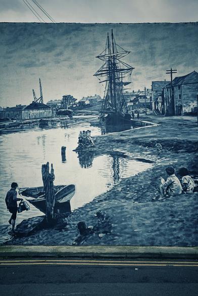 A monochrome mural depicting a historical waterfront scene. In the foreground, two children are on a grassy bank, one looking towards the water, and another boy is by a small boat. The mural features a large sailing ship docked in the water, with other boats and waterfront buildings visible in the background. The overall tone is reminiscent of an old photograph or painting, with a blueish tint.