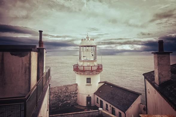 A striking view of a white lighthouse with a red railing, standing prominently on a rugged coastline under a dramatically cloudy sky. The dark, choppy sea stretches to the horizon. Parts of two buildings with dark roofs are visible on either side of the lighthouse, framing the scene.
