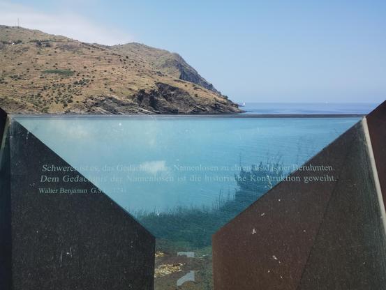 Blick auf eine Glasscheibe mit der Aufschrift:
»Schwerer ist es, das Gedächtnis der Namenlosen zu ehren als das der Berühmten.
Dem Gedächtnis der Namenlosen ist die historische Konstruktion geweiht.
Walter Benjamin, G.S. I (auf der Glasscheibe als L), 1241«
Im Hintergrund Meer und eine Küste