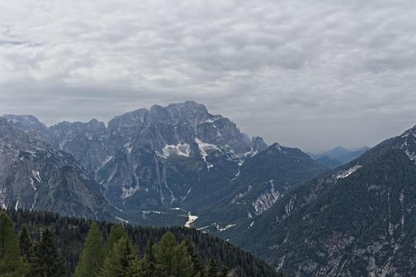This photo captures a majestic mountain range under a heavily overcast sky in the Julian Alps, from the perspective of Mount Lussari. Jagged peaks rise dramatically, layered with dark stone and scattered patches of snow or scree. Pine-covered slopes descend into a deep, forested valley, which is bisected by a pale, winding riverbed or road. The sky above is filled with thick, textured clouds — light grey, with subtle shadowing that hints at depth and movement, but no direct sunlight. The mood is calm, slightly somber, and natural. The landscape feels vast and remote, with cool tones and strong contrasts between the rugged mountains and the soft, clouded sky.
