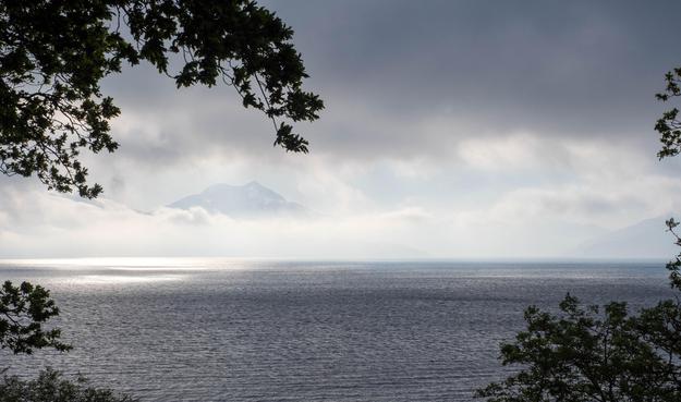 Distant hills across the sea, emerging from a morning mist. The sun illuminates a patch of sea through a gap in high dark cloud.