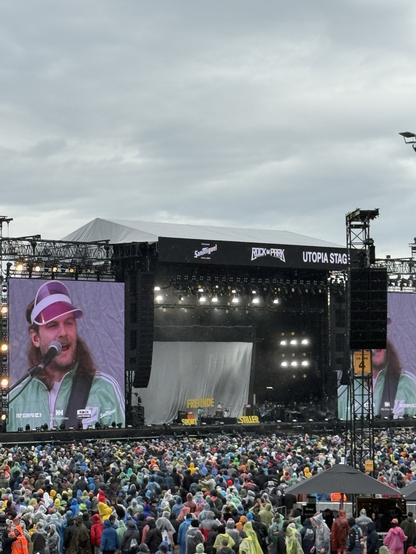 A musician performing on stage during a concert at the Utopia Stage, with a large audience visible wearing colorful raincoats. The scene is set against a cloudy sky.