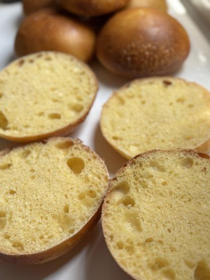 A close-up view of freshly baked sourdough rolls, cut in half to reveal a soft, airy interior with small holes. In the background, whole rolls are visible.