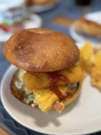 A close-up of a sourdough burger featuring a golden-brown bun, crispy fish fillet, egg, avocado and ketchup. The burger is served on a white plate with a partially visible breakfast item in the background.