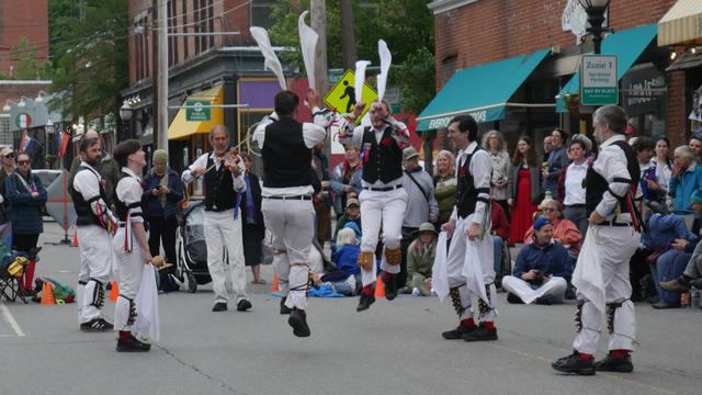 Six morris dancers with white shirts and trousers and black vests, decorated with multicolor ribbons, dancing in a street. Four are standing and two are in motion, feet off the ground and hankies in both hands raised. Behind them is a musician playing a fiddle and an audience including some other morris dancers.