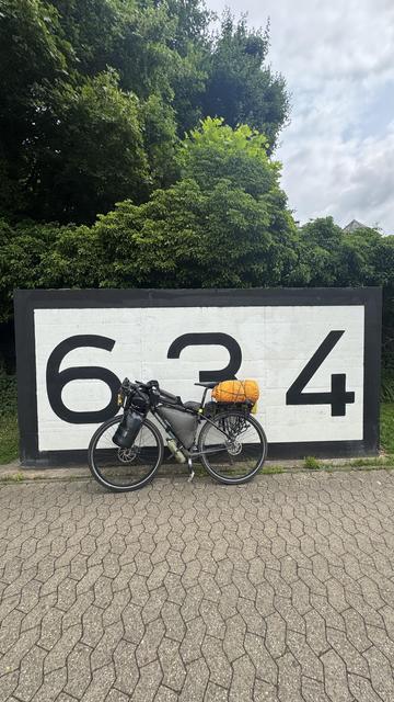 Ein Fahrrad ist neben einer großen Wand geparkt, die mit den Zahlen "634" in fetter, schwarzer Schrift bemalt ist. Im Hintergrund ist üppiges Grün zu sehen, und der Boden verfügt über einen gemusterten Bürgersteig.