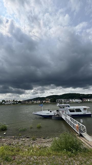 Eine Fähre namens "NIXE" liegt an einem Dock entlang eines Flusses fest, umgeben von grünem Laub und felsigem Gelände. Der Himmel ist mit dunklen Wolken bedeckt, während Gebäude im Hintergrund am gegenüberliegenden Ufer sichtbar sind.