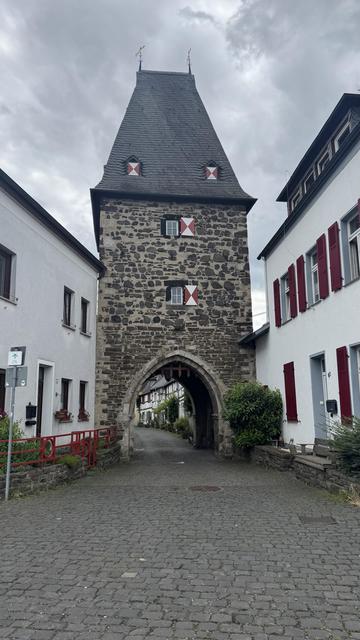 Eine schmale Kopfsteinpflasterstraße, flankiert von weißen Gebäuden, führt zu einem historischen Steinturm mit einem spitzen Dach und dekorativen Fensterläden. Der Turm verfügt über einen gewölbten Eingang, und die Szene spielt unter einem bewölkten Himmel.