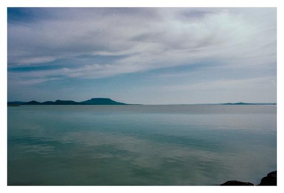 Color photograph of Lake Balaton. The lake is deserted in this detail, there are no boats or swimmers to be seen. The picture is very blue. A few clouds can be seen in the sky. A few stones from the shore are cropped in the foreground at the bottom right. In the background you can see the mountain ranges of the typical volcanoes.