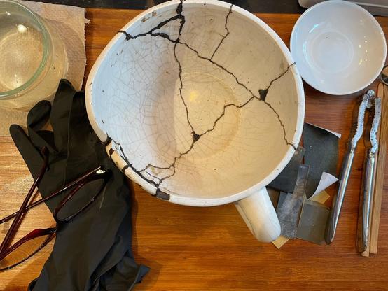 Foto eines bereits mit Urushi geklebten Keramiktopfes, der weiterbearbeitet werden soll. Daneben Handschuhe, Lesebrille, ein niedriges Glas mit Wasser, Schleifpapier und eine flache Schale für den Urushi-Lack