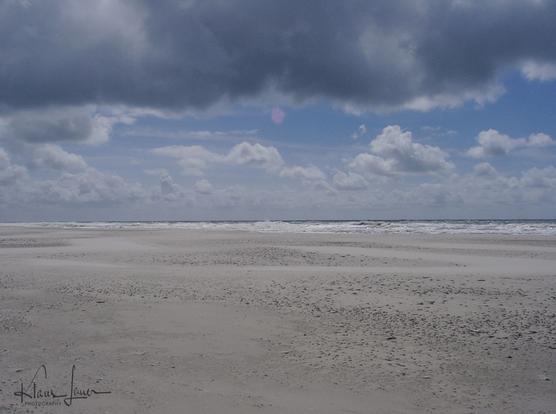 Auf dem Foto sieht man den flachen Sandstrand des Kniepsandes, einer Amrum vorgelagerten Sandbank. Die kabbeligen Wellen der Nordsee laufen auf den Strand auf. Der blaue Himmel ist mit weißen und grauen Wolkenfetzen gesprenkelt.