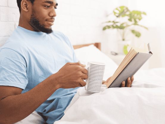 Ein junger Mann sitzt im Bett mit einer Tasse und liest ein Buch