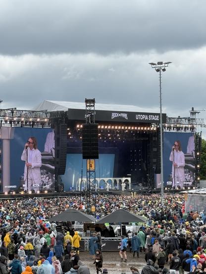 A performer in white attire stands on the Utopia Stage at a festival, with large screens displaying them. The audience, wearing colorful raincoats, is gathered in front of the stage, under a cloudy sky. The scene conveys a vibrant yet rainy