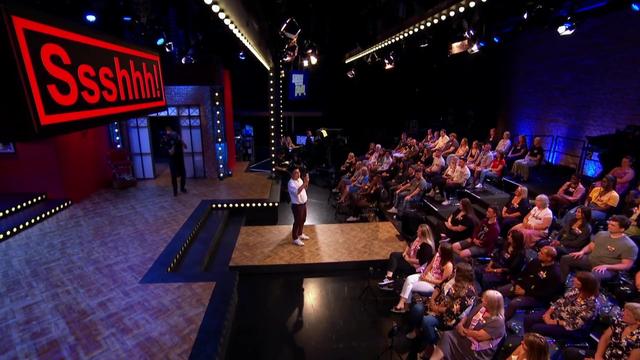A side view of a comedian on stage, facing the audience, and a large "Sssssssh!" sign lit up red.