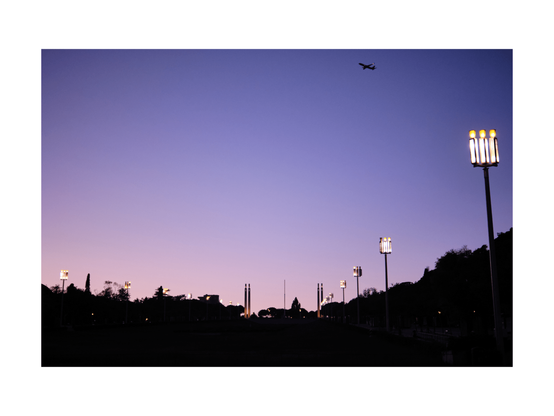 violet gradient sky over a black silhouette of an urban park situation with shinig street lights left an right aligned. in top right corner an aeroplane