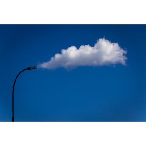 A photo showing the top curve of a streetlamp, with a white cloud drifting across the sky in the background, creating the illusion that the cloud is coming out of the steeetlamp. This is seen with a deep blue skye behind it.