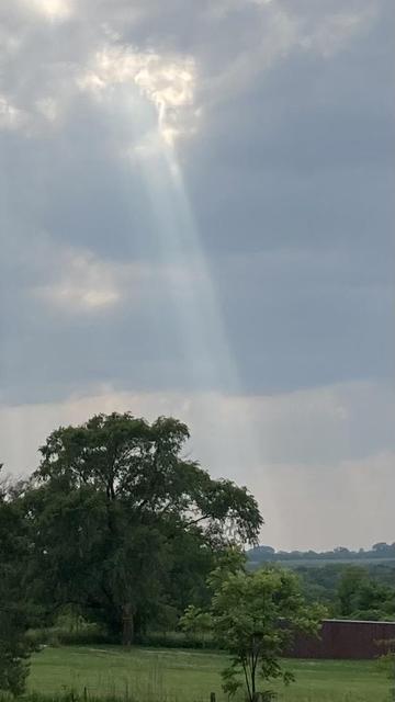 A sunbeam pierces the gray and white clouds and shines down on the extremely green fields and trees.