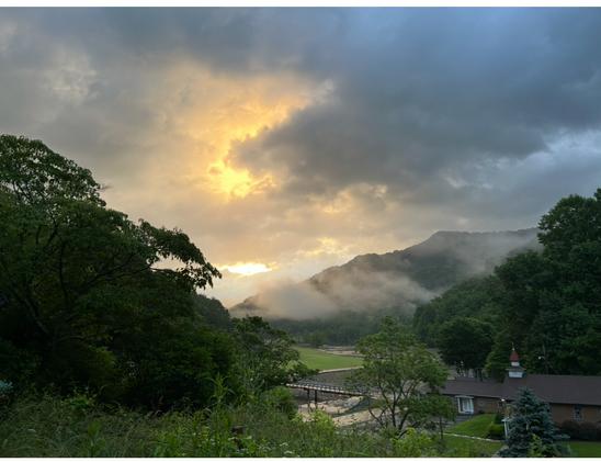 A dramatic sunrise graces a dark grey blue clouded sky above, with a centered splash of gold light breaking through, dramatically highlighting them in brilliant outline. Soft rays penetrate wisps of mist and lend white tracery emanating in all directions. A sloping mountain range ascends diagonally center left to right in the background. A small diamond patch of green meadow sits just at center above an overgrown field of green, with trees in full leaf bordering right and left. The rooftop of a small chapel in dark red/brown hues is sited bottom right, with a hint of river and road showing just to its left