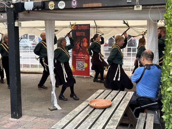 North west style Morris dancers wearing green and black, dancing in front of a Red Cuthbert Morris roll out banner