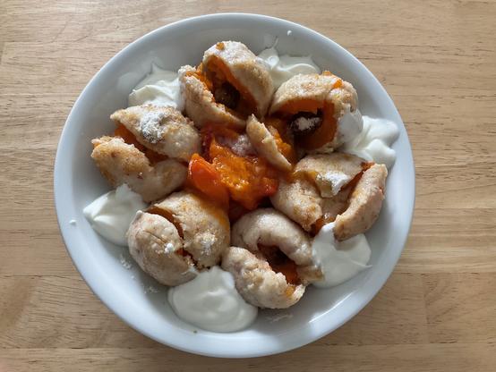 6 schon geöffnete, in BröselButter geschwenkte Aprikösenklöße mit Quark und Puderzucker, in der Mitte noch AprikosenRöster - alles in einem weißen Teller auf einem hellen Holztisch