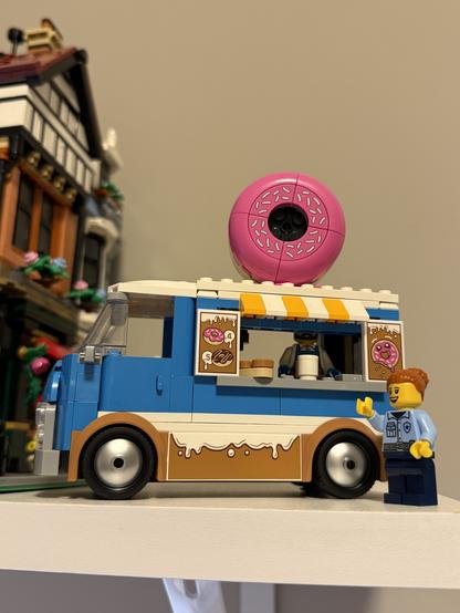 Lego donut truck model with a large pink donut on top. A smiling Lego policewoman stands beside it. Nearby, a detailed Lego building. Playful and cheerful.