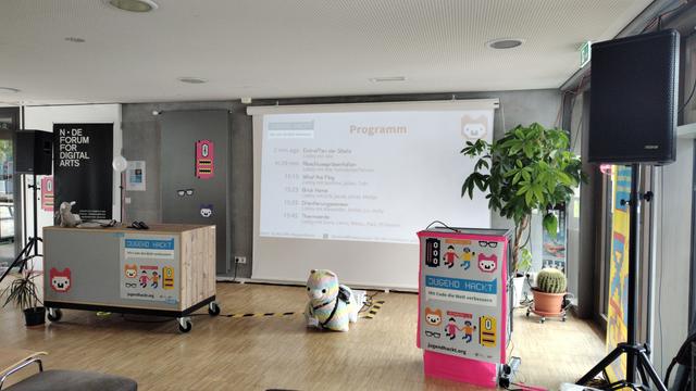 Zwei Rednerpulte, eine Leinwand, ein riesiges Plüschalpaka, in einer Ecke eines Raums, die zur Bühne umfunktioniert wurde.