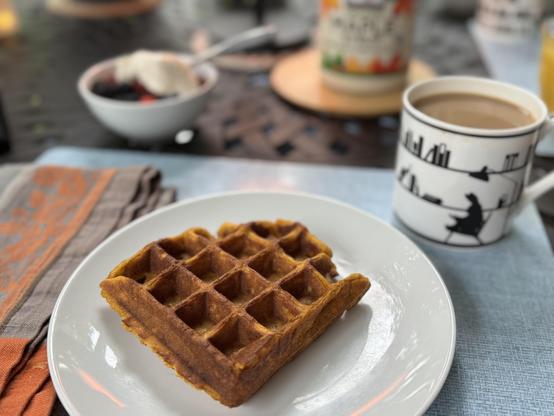 A deep-orange Belgian pumpkin waffle rests on a small white plate at a breakfast table.  The plate sits on a blue tableboard, which also has a napkin, and a Ham rabbit coffee mug (the Reader.)  Behind and out of focus is a small bowl of berries with yogurt, and a coaster holding a large jug of Kirkland maple syrup.  

The table itself is a black metal trellis table, upon which a variety of other objects rest, which are mostly all out of focus and barely seen;  (another place setting, a small lamp, 2 plants.)

The Man has exceeded excellence on Sunday morning breakfast yet again.
