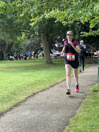Fiona running and smiling in amongst leaves and greenery during Magna Carta marathon. Fiona wears a magenta top on a cap and the race number 28, in shorts and fluorescent pink trainers.