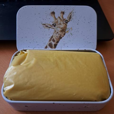 Eine kleine offene Metallbox mit einem gefaltenen gelben Sack darin. Am Deckel, welcher dahinter lehnt, ist eine gezeichnete Giraffe die Blumen mampft.