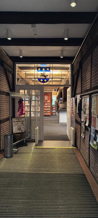 Entree met balken plafond, vakwerk muren en glas in lood boven de tussendoor met wapen en jaartallen 1730 en 1980. Door de openstaande tussendeur is de bibliotheek te zien.