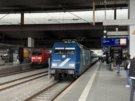 Picture at a station with platforms left and right. In the middle and middle left two trains are visible. One with a red front and other on the right with a blue front.