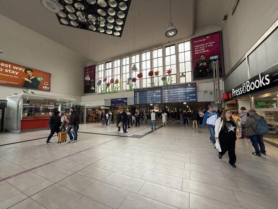 Inside a large hallway cq entrance of the Düsseldorf Hbf station. Some people left and right. Big windows up ahead. On the right “Press&Books” and up ahead also a departure sign.