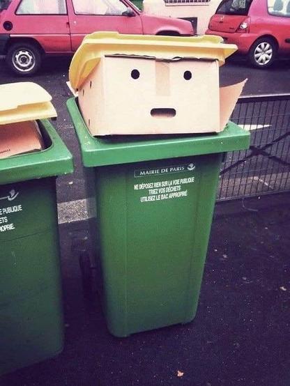 A green recycling bin in Paris humorously transformed to resemble a cartoonish human face using a cardboard box with cut-out eyes and mouth. A yellow lid positioned like hair gives it a strong resemblance to Donald Trump’s iconic hairstyle. The scene is in an urban street with parked cars in the background. The label on the bin reads “Mairie de Paris.”