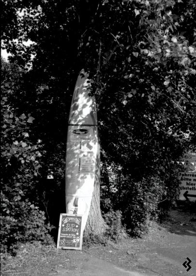 Black and white photo of a surfboard leaning on a tree. Pieces of paper with offers of the nearby lakeside café / boat rental are attached to the surf board. On the ground, there is a little blackboard leaning on the surfboard with the text "SUP Station" on it