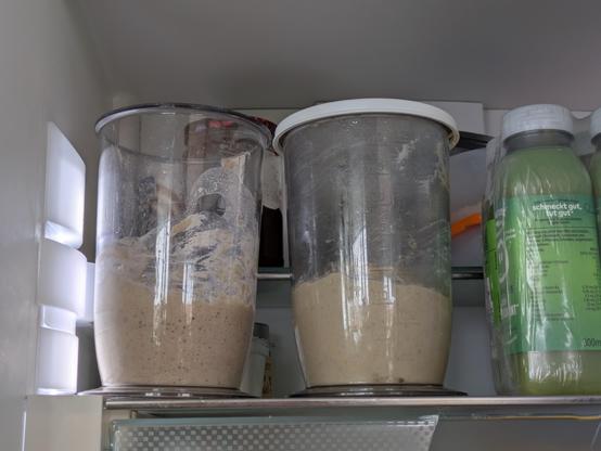 Das Bild zeigt den Innenraum einer Kühlschranktür. Auf der oberen Regalebene stehen zwei transparente, große Behälter mit Deckel, die mit einer braunen, brotähnlichen Masse gefüllt sind. Die Masse hat eine leicht feste Textur und enthält kleine Luftblasen, was darauf hindeutet, dass sie aktiv fermentiert wird. Der Behälter auf der linken Seite hat einen teilweise abgenutzten Deckel, während der auf der rechten Seite einen sauberen Deckel trägt.