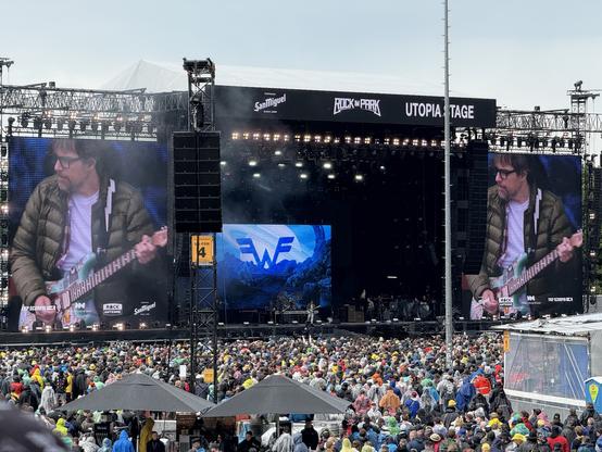 A musician playing guitar on stage at a festival, with a large screen displaying their image. The audience, wearing ponchos, is gathered in front of the Utopia Stage at Rock im Park.