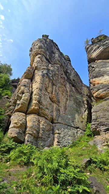 Eine massive, hohe Felswand aus geschichtetem Sandstein, von unten betrachtet, mit grünem Bewuchs am Fuße und unter blauem Himmel.