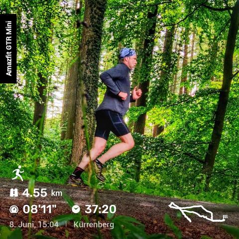 Ein Mann joggt auf einem Waldweg, umgeben von üppigem Grün. Er trägt ein graues Langarmshirt, schwarze Shorts und schwarze Laufschuhe. Ein blaues Stirnband hält sein Haar zurück. Links im Bild steht der Schriftzug „Amazfit GTR Mini“. Unten werden Laufdaten angezeigt: zurückgelegte Strecke von 4,55 km, eine Pace von 08’11“, eine Gesamtzeit von 37:20 Minuten. Das Datum ist der 8. Juni, 15:04 Uhr, und der Ort ist Kürrenberg. Rechts unten ist eine Karte mit der gelaufenen Strecke zu sehen.