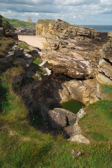 A coastal scene at Rumbling Kern, Howick, Northumberland, England, featuring rugged sandstone cliffs and rocky formations under a partly cloudy sky. The foreground is dominated by green grassy slopes dotted with small pink flowers, which lead down to a rock pool filled with green water nestled among weathered boulders. Beyond this, flat sandstone platforms extend toward a sandy beach enclosed by taller cliffs with patches of grass on top. In the distance, a solitary stone house stands near the edge of the cliff, overlooking the calm blue sea on the horizon, creating a striking contrast between natural ruggedness and human habitation.