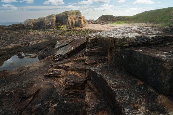 A rugged coastal scene at Rumbling Kern, Howick, Northumberland, England. In the foreground, dark, layered sedimentary rocks with rich brown and black hues are sharply defined, revealing intricate textures and natural striations. Further back, the rocky shore extends towards a cluster of large, weathered sandstone cliffs with patches of green moss and lichen. To the right, the terrain rises gently with grassy slopes leading to sandy patches, under a partly cloudy sky with soft, diffused sunlight illuminating the landscape, creating a serene yet wild coastal atmosphere.