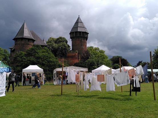 Der Bergfried in der Bildmitte, dahinter mächtige Wolken am Himmel. Davor die Wiese im Sonnenschein. Auf Wäscheleinen hängt Wäsche, die heute voraussichtlich nicht trocken werden wird.