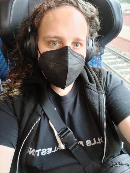 A selfie of Mirah in a train window seat. She's wearing a black t-shirt that reads "Dolls for Palestine," an open black sleeveless hoodie, headphones, and a black mask